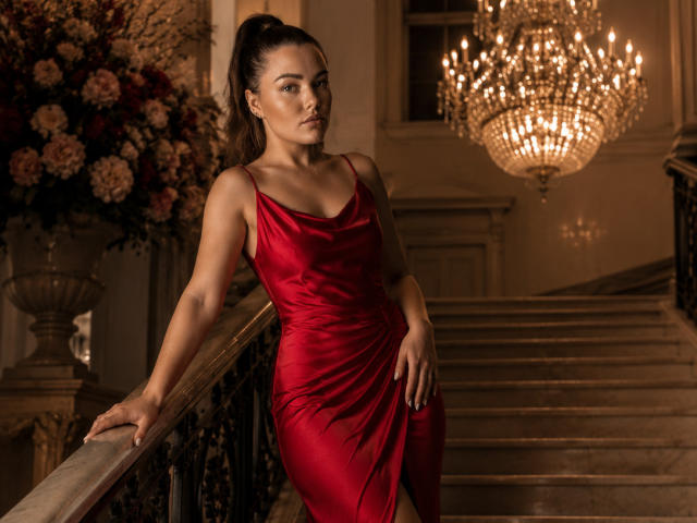 Sansarra with toned arms, wearing a red satin dress, leaning on a staircase, her ass slightly raised, dark hair in a ponytail, chandelier above, flowers to the left.