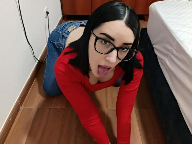 Devorah69 on all fours, tongue out, wearing glasses, red long-sleeve top, blue jeans, black hair, wooden floor, bed on right, wooden cabinet in background, looking up.