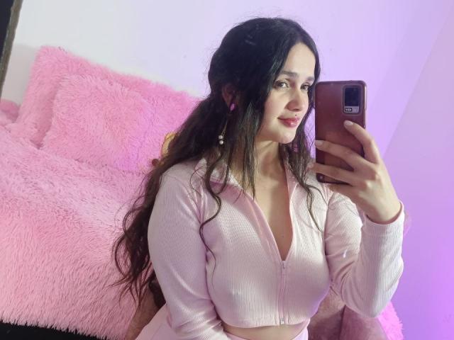 NataliaRivas, con lunghi capelli scuri, indossa una maglietta rosa con zip, tiene in mano un telefono, ci sono cuscini rosa e soffici, sta scattando un selfie allo specchio, mostrando il seno, indossa orecchini rosa, rossetto rosa, sta sorridendo, l'illuminazione è viola..