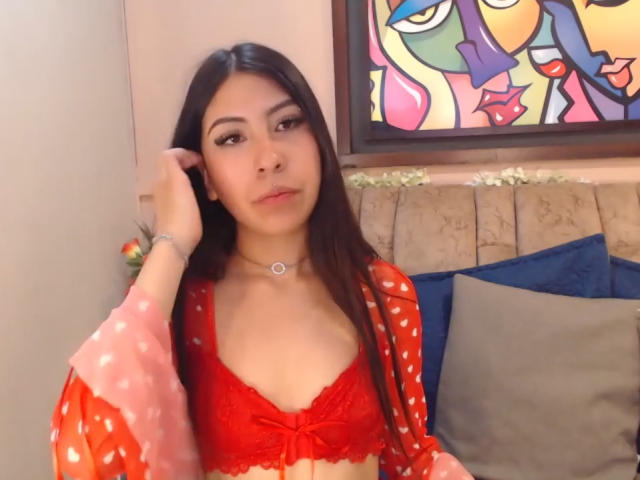LinaSpearce with long dark hair, wearing a red lace bralette and pink polka dot robe, chomping on her right hand, necklace, and blue pillows in the background.