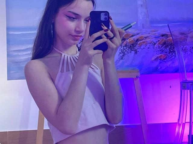 MimiPreaty mit langen braunen Haaren, pinkem Eyeliner und Reifenohrringen, trägt ein pinkes Halstopp und macht ein Selfie mit einem schwarzen Handy, auf dem ihre kleinen Brüste zu sehen sind..