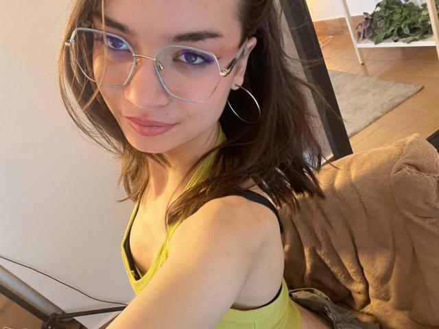 MimiPreaty, avec des lunettes, des boucles d'oreilles pendantes et un débardeur jaune qui dévoile ses seins, souriant à la caméra, avec des cheveux bruns, du fard à paupières violet et un joli visage..