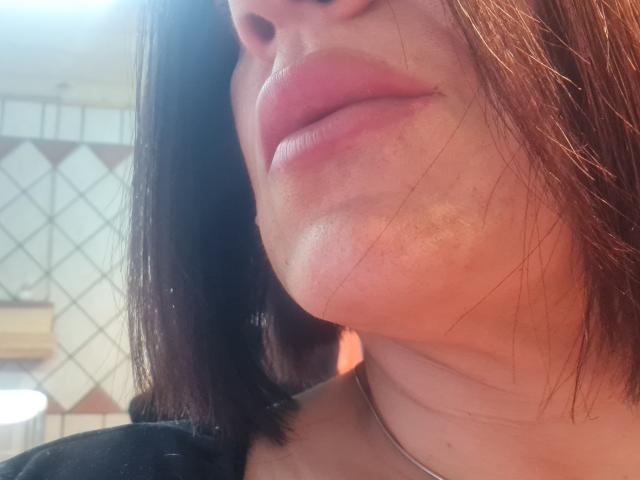 Les lèvres de Pantera69X sont légèrement retroussées, sa peau a un éclat subtil et elle a les cheveux bruns, mi-longs. Son menton est légèrement relevé, et on peut voir un collier en argent..