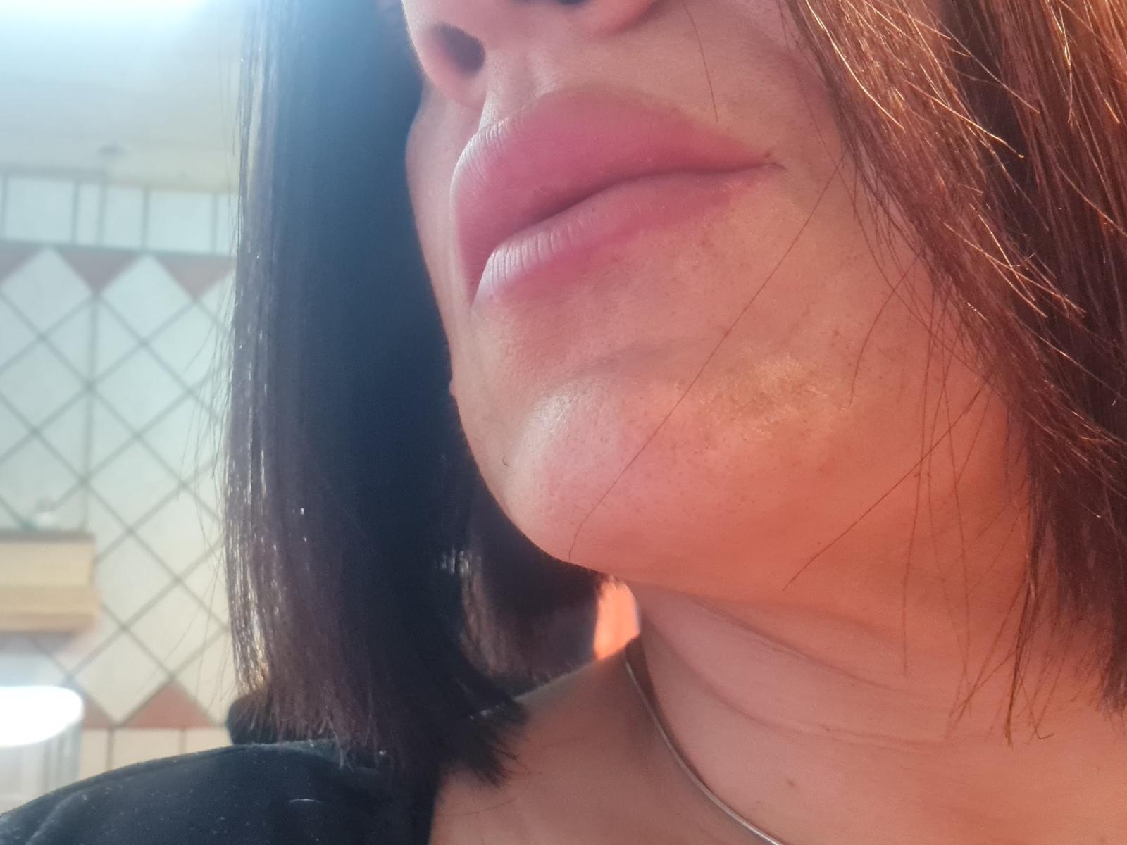Os lábios de Pantera69X estão levemente curvados, com um brilho sutil na pele e cabelos castanhos na altura dos ombros. Seu queixo está ligeiramente levantado, mostrando um colar de prata..