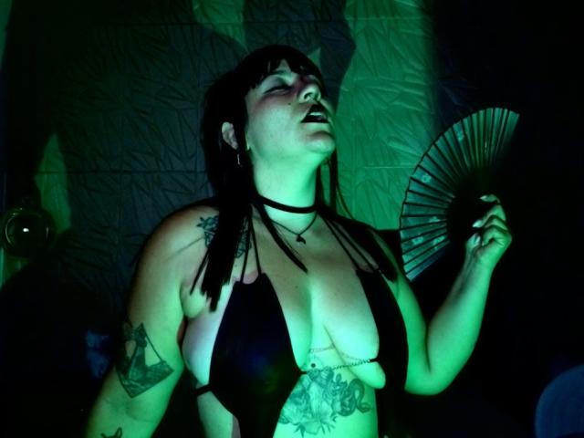 NaniSky with green-tinted skin, black bikini, large tits, tattoos, holding fan, dark hair, bangs, choker, eyes closed, mouth open, standing, shadowy background.