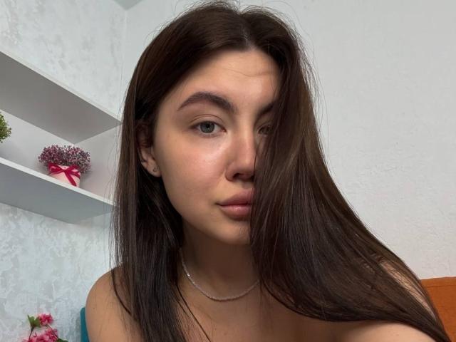 InesGueningsman with long brown hair, light skin, and small tits, wearing a necklace, looking at the camera with a neutral expression. Shelves with pink flowers in the background.