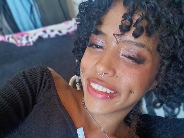 LunaKeller heeft krullend zwart haar, bruine huid, neuspiercings en lipiercings, met roze oogschaduw, knippert, glimlacht, draagt een zwarte top en een ketting..