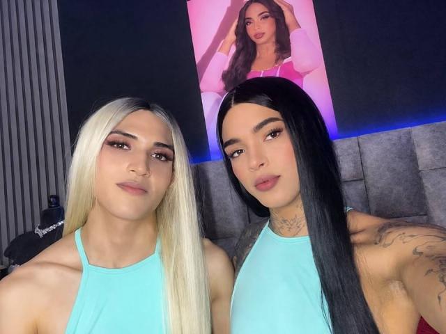 MelodyCamilaTS with long blonde and black hair, wearing teal tops, showing tattoos, and looking at camera, standing in front of a pink portrait of another MelodyCamilaTS.