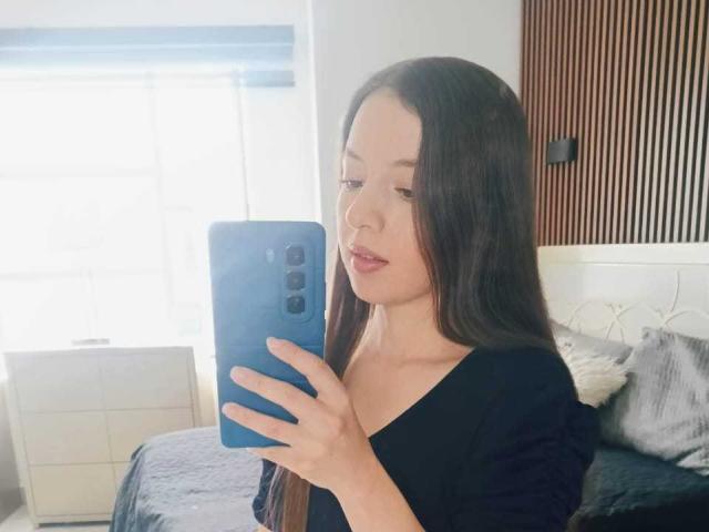 VioletaNova with long dark hair, wearing a black top, holding a blue phone, taking a mirror selfie, showing subtle pink lipstick and a calm expression.
