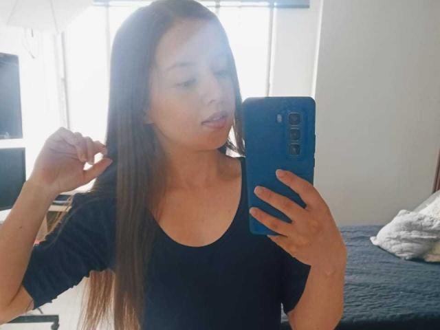 VioletaNova with long brown hair, black tee, pouty lips, holding blue phone, mirror selfie, slight smile, right hand touching hair, bed with gray blanket in background.
