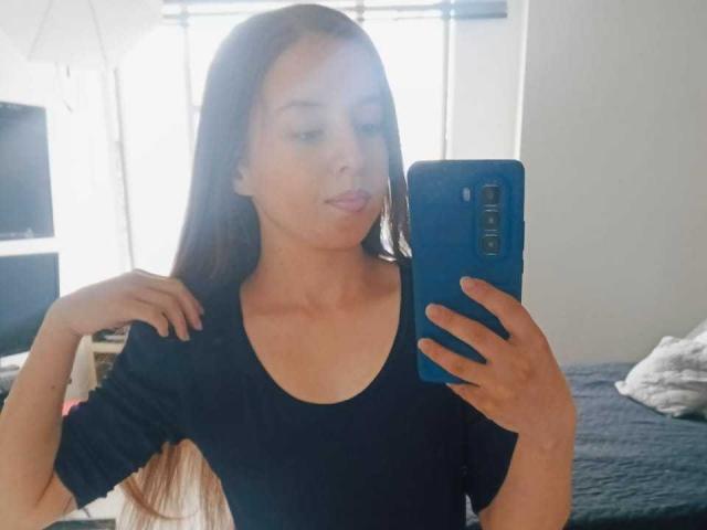 VioletaNova with long brown hair, wearing a black top, taking a selfie in a mirror with a blue phone, showing a cute pout, her hand on her shoulder.