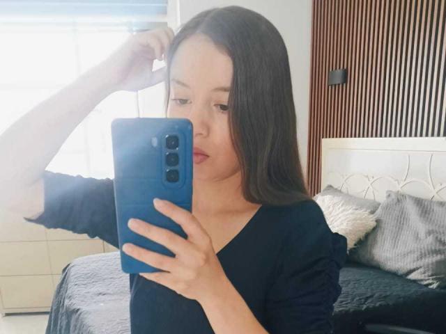 VioletaNova with long brown hair, wearing a black shirt, holding a blue phone, taking a mirror selfie, her arm raised, light in her face, red lipstick, bedroom setting.
