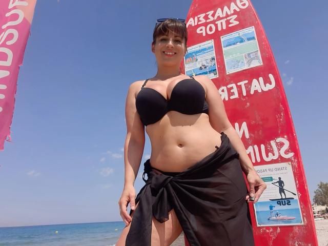 LaLeticia, sensuelle, avec un soutien-gorge noir et une jupe noire nouée à la taille, se tient sur la plage, montrant sa poitrine, son petit ventre, et sourit. Elle porte des lunettes de soleil sur la tête. Le ciel est bleu, et on voit l'océan..