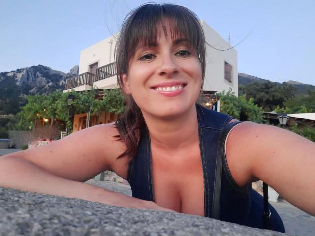 LaLeticia, con cabello castaño y flequillo, sonriendo a la cámara, vestida con una camiseta vaquera sin mangas que deja ver sus pechos, apoyada en una superficie de piedra, al aire libre en un entorno montañoso..