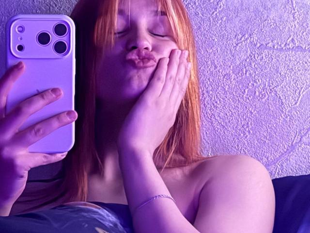 Cheri Worstell, avec ses cheveux roux, les lèvres pincées, une main sur son visage, prend un selfie avec un iPhone violet. Son épaule est nue, et elle porte un bracelet en argent..