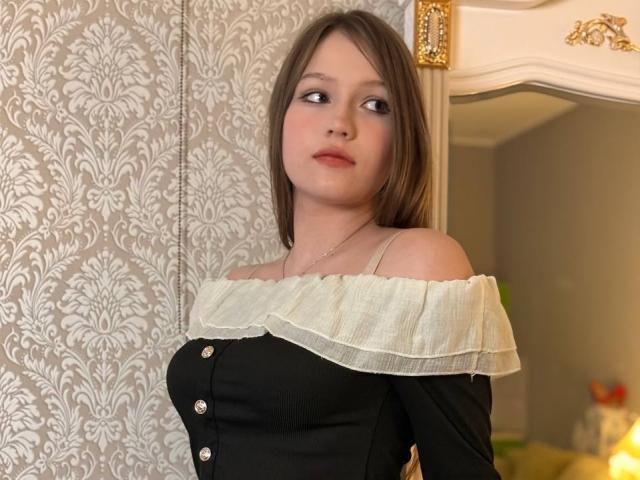 Bubochka with light brown hair, off-shoulder black top with white ruffle, small tits, buttoned down, looking to the side, in a softly lit room.