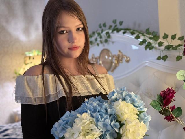 Bubochka with long brown hair, off-shoulder black and white dress, holding blue and white hydrangea bouquet, sitting on white, ornate bed with green plant in background.