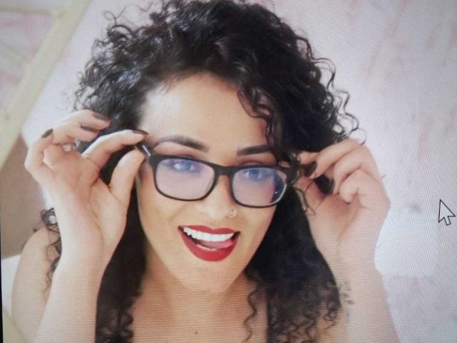 AliciaShadow mit lockigem, schwarzem Haar, blauen Augen, rotem Lippenstift und einer schwarzen Brille, die sie mit beiden Händen hält, lächelt mit offenem Mund und zeigt ihre Zähne..