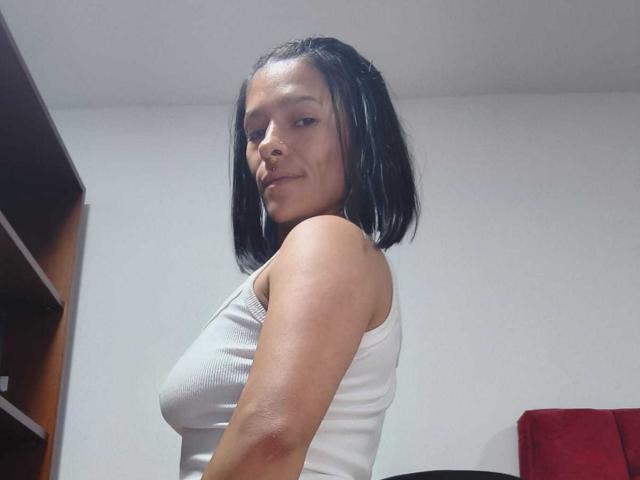 AmelieBlake with medium tits, wearing a white tank top, has black hair in a bob, looking at the camera with a slight smirk, in a simple room.