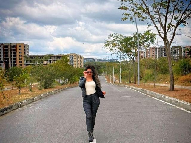 AntonelaRiveiro with long black hair, wearing red sunglasses, white top, black puffy jacket, and black pants, standing on a deserted road, holding a phone to her ear.