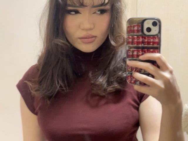 DebPlagens, avec ses cheveux bruns ondulés, portant un pull rouge à col roulé, prend un selfie avec un téléphone dans un miroir. Ses seins sont visibles sous le haut moulant..
