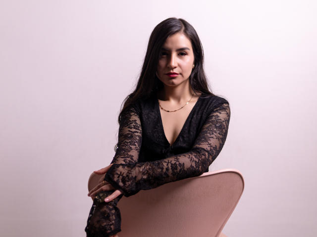 Jade Harpper, con lunghi capelli neri, indossa un abito nero in pizzo, è seduta su una sedia rosa, con le braccia incrociate, un sorriso appena accennato, pelle chiara, una piccola collana d'oro, rossetto rosso..