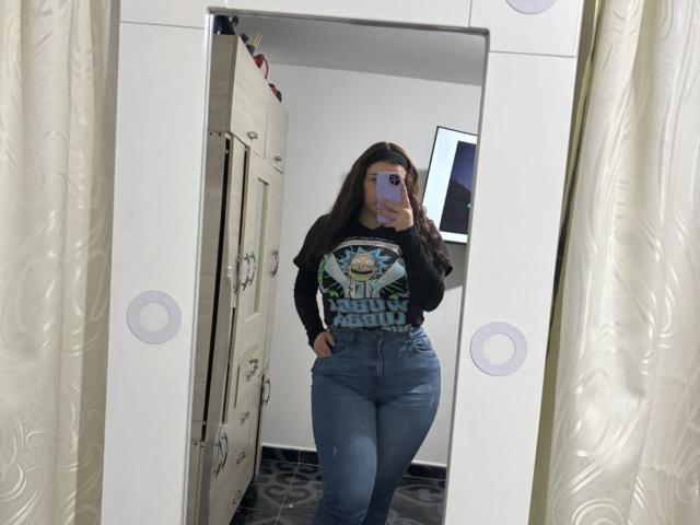 VenusxMusa met lang bruin haar, een zwart T-shirt met een grafisch ontwerp en een blauwe spijkerbroek met hoge taille, maakt een foto van zichzelf in de spiegel, waarbij haar ronde billen en kleine borsten te zien zijn, in een kleedkamer..