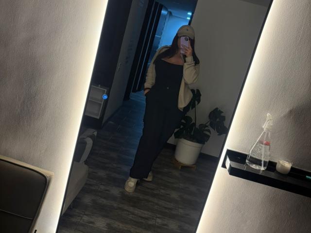 VenusxMusa vestida con pantalones y camiseta negros, una chaqueta beige y un sombrero, se toma un selfie con el teléfono, mostrando sus glúteos con zapatillas blancas, en una habitación con poca luz..