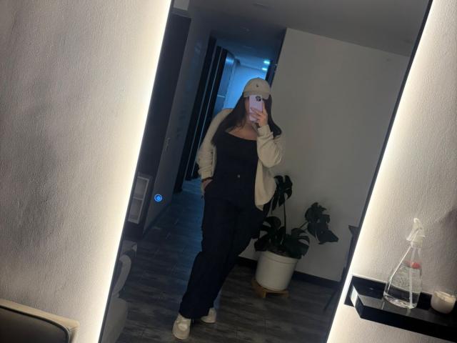 VenusxMusa vestindo roupa preta, tênis brancos, jaqueta bege, boné branco, segurando um celular, selfie no espelho, mão na cintura, planta em vaso, piso de madeira escura, pequena garrafa de água, iluminação fraca..
