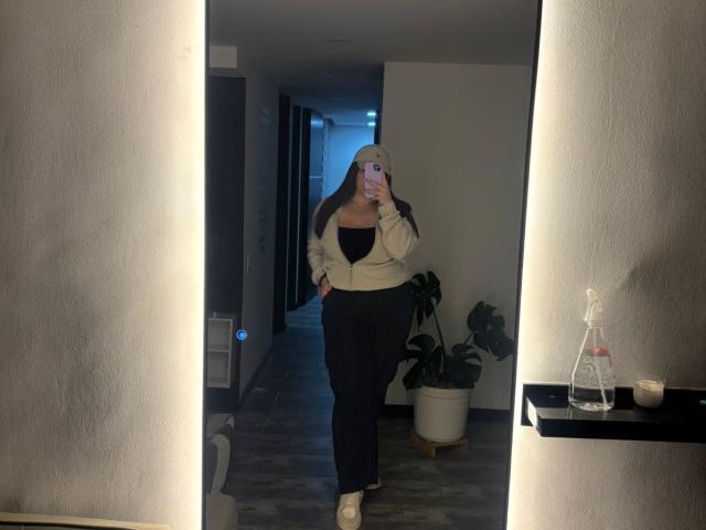 VenusxMusa in een spiegel-selfie, met een beige muts, een witte jas, een zwarte top, zwarte broek, witte sokken, een telefoon vasthoudend, staand in een zwak verlichte kamer..
