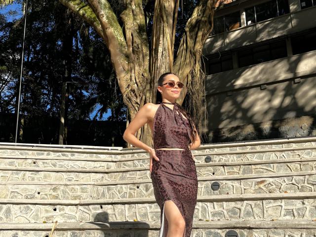 Alana Blake steht vor einer Treppe, trägt ein trägerloses, dunkelbraunes Kleid mit Leopardenmuster und einem Schlitz am Oberschenkel, hat die Hände in den Taschen, lächelt und trägt eine Sonnenbrille. Im Hintergrund ist ein Baum zu sehen..