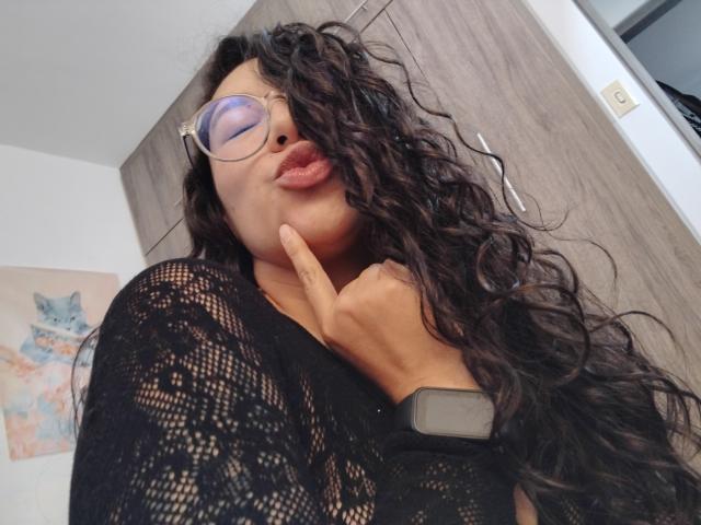 Mllevv, con capelli neri ricci, occhiali e labbra piene, che tocca il mento con un dito, indossa una camicetta in pizzo e uno smartwatch, ha labbra rosa gonfie, pelle marrone chiaro, è sexy e sicura di sé..