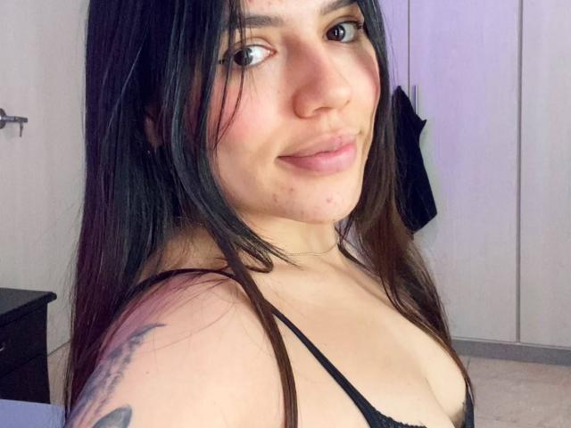 SophiaMarx with long black hair, brown eyes, and light skin, smiling slightly, showing small tattoos on her right shoulder, wearing a black spaghetti strap top, revealing her small tits.