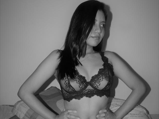 LolaGibson in black lace bra, hands on hips, looking to the side, medium tits, dark straight hair, white wall, bed with pillow in background, black and white photo.