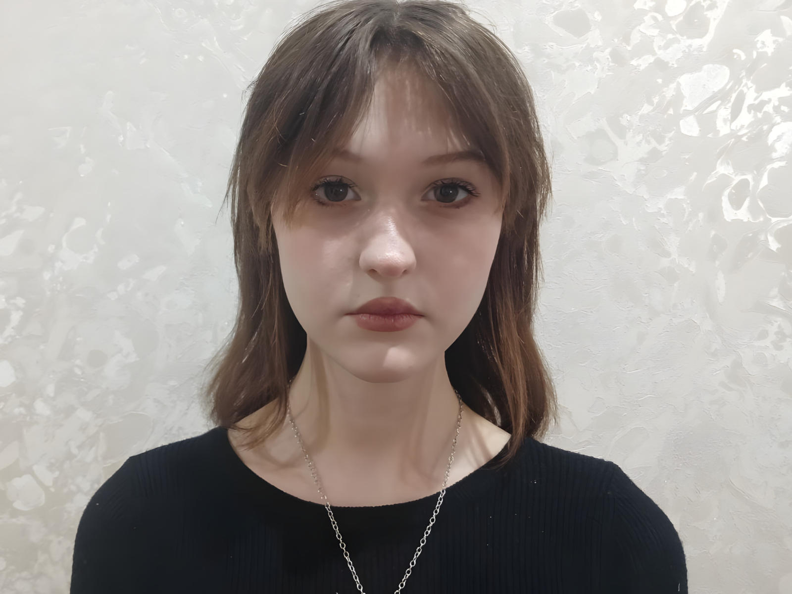 LillyDupass has pale skin, brown eyes, and straight brown hair with bangs. She's wearing a black rib-knit top and a silver necklace. Her expression is neutral.