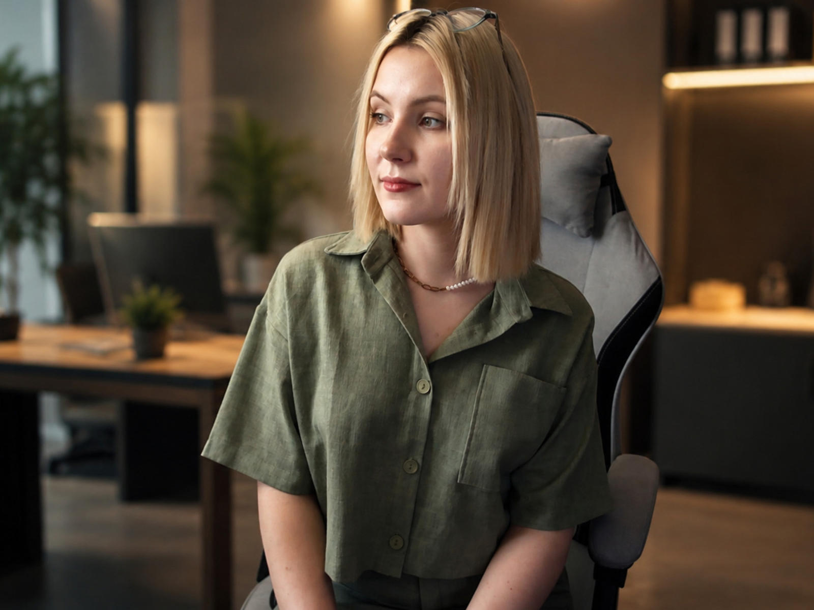 Lidia Ockimey, con caschetto biondo, camicia verde a bottoni, seno piccolo, seduta su una sedia da ufficio, con un leggero sorriso, occhiali sulla testa, collana, illuminazione soffusa, sfondo sfocato, espressione calma, rilassata..