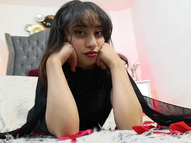 CamiVargas, com cabelo preto, batom vermelho e blusa preta transparente, deitada na cama com as mãos no queixo, cercada por pétalas vermelhas, olhando para a câmera com uma expressão sedutora..