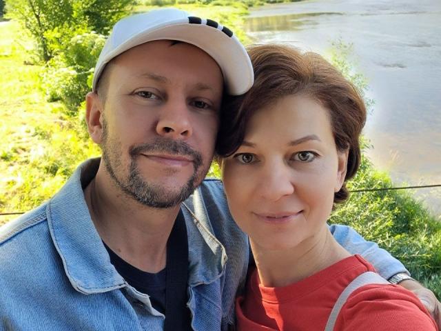 HotDuoLove, ein Mann mit Bart, einer hellblauen Mütze und einer Jeansjacke, und eine Frau mit kurzen, braunen Haaren und einem roten Hemd, stehen eng beieinander und lächeln in die Kamera..