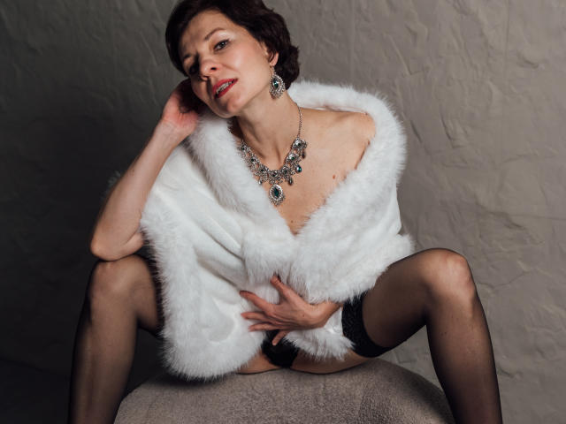 HotDuoLove mit kurzen, braunen Haaren, trägt einen weißen Pelzmantel, schwarze Spitzenstrümpfe und eine dicke silberne Halskette, sitzt mit gespreizten Beinen, eine Hand auf dem Mantel, und wirkt verführerisch..