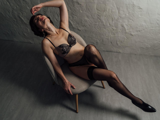 HotDuoLove è una donna attraente con piccoli seni, che indossa un reggiseno di pizzo nero, biancheria intima nera e collant neri, seduta su una sedia grigia, con gli occhi chiusi, un braccio sollevato e la testa reclinata all'indietro..
