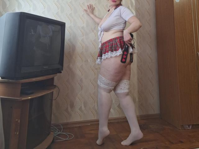 Sabrina, avec sa peau pâle, ses petites seins et ses hanches rebondies, porte un crop top blanc, une jupe rouge à carreaux, des bas blancs en dentelle et tient une pagaie noire. Elle se tient dans une pièce..