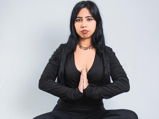 AyameFoxy, com cabelo preto, vestindo uma camisa preta com decote profundo em V, mãos em posição de oração, sentada com as pernas afastadas, usando um colar de contas pretas, olhando diretamente para a câmera..
