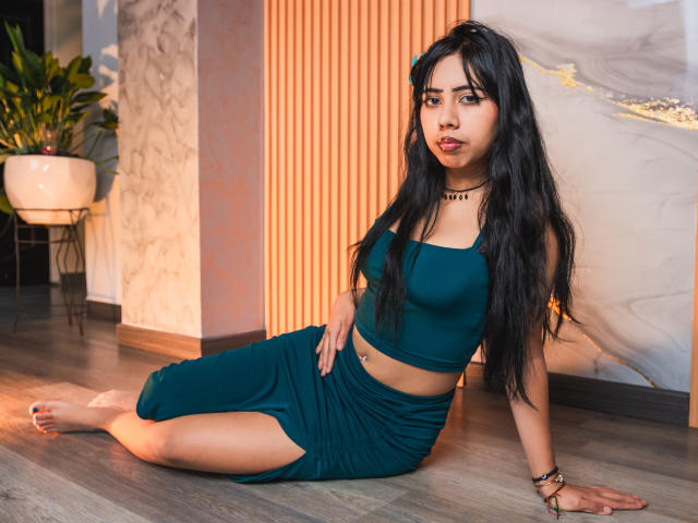 AyameFoxy sitzt auf dem Holzboden, trägt ein türkisfarbenes Crop-Top und einen passenden Rock, hat langes, schwarzes Haar, eine Choker-Kette, ist barfuß, schaut in die Kamera, hat einen kleinen Bauchnabelpiercing und dezentes Make-up..