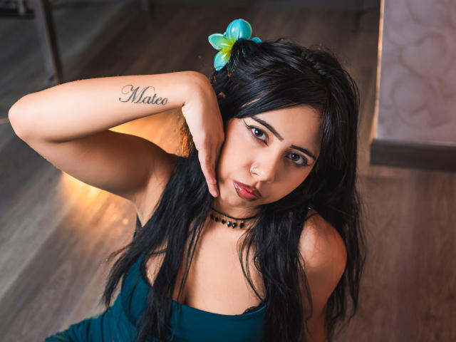 AyameFoxy mit schwarzen Haaren, einer blauen Blume und einem grünen Kleid, die ihre Nase berührt, ihre Brüste zeigt und ein "Mateo"-Tattoo an ihrem Handgelenk trägt, und eine schwarze Perlenkette trägt..