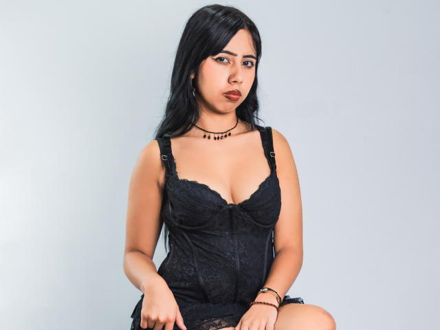 AyameFoxy mit langen, schwarzen Haaren, trägt ein schwarzes Spitzen-Dessous-Kleid, zeigt ihre Brüste, hat braune Augen, roten Lippenstift, eine schwarze Halskette und ein Armband und sitzt vor einem schlichten Hintergrund..