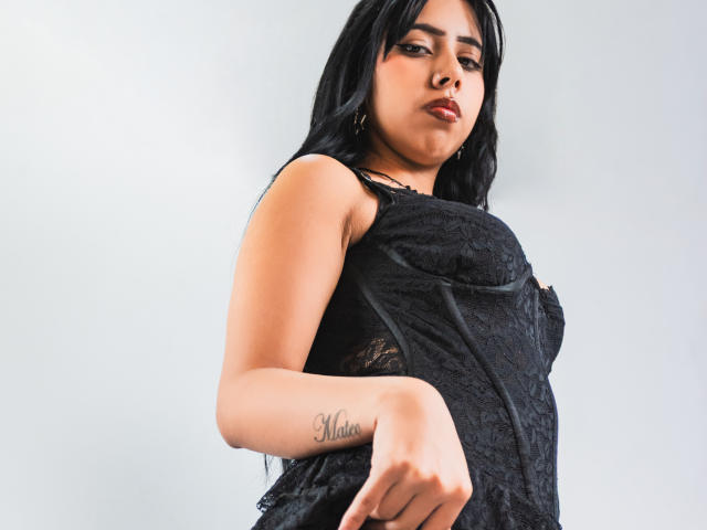AyameFoxy mit langen, schwarzen Haaren, trägt eine schwarze Spitzenbluse, auf deren der rechten Unterarm ein Tattoo zu sehen ist, das "Mata" sagt. Ihre Brüste sind leicht durch die Spitze hindurch sichtbar..