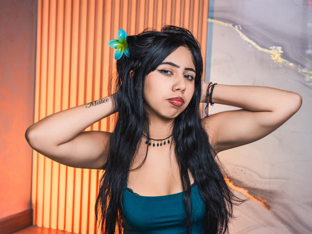 AyameFoxy met lang zwart haar, een blauwe bloem, draagt een turquoise top, armen omhoog, handen achter het hoofd, kijkt naar de camera, rode lipstick, een zwarte kralenketting, tatoeages op de rechterarm..
