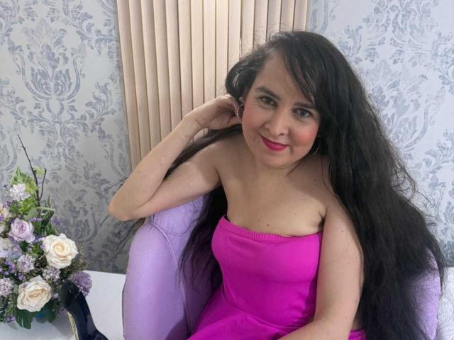 Diana Lane mit langen schwarzen Haaren, einem pinkfarbenen Kleid ohne Träger und rotem Lippenstift, sitzt auf einem violetten Stuhl, lächelt, hat eine Hand im Haar, auf einem Tisch steht ein Blumenstrauß mit Rosen und anderen Blumen..