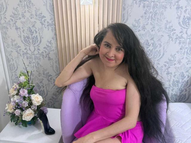 Diana Lane con lunghi capelli neri, vestito rosa senza spalline, seduta su una sedia viola, sorridente, con una mano sulla testa, rossetto rosa, parete bianca, vaso di fiori e scarpa nera in primo piano..