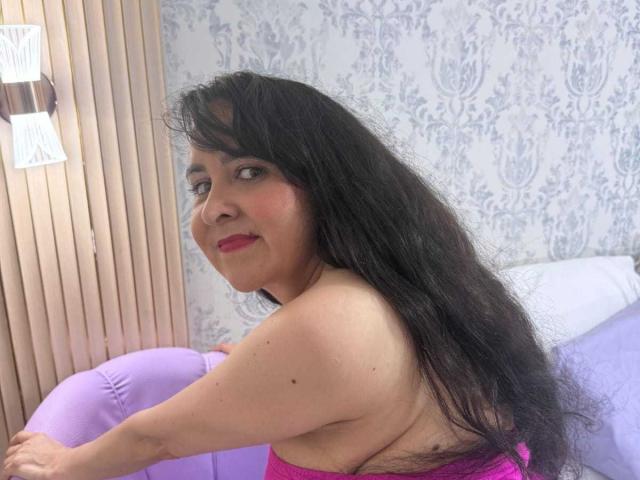 Diana Lane mit langem, schwarzem Haar, rosa Lippenstift und rotem Rouge, trägt ein rosa Oberteil und sitzt auf einem violetten Stuhl, wobei ihre nackten Schultern und ein Teil ihrer Brust sichtbar sind..